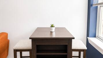 Una habitación con una mesa de madera, dos sillas, una planta en maceta, un reloj de pared y un ventanal con persianas.