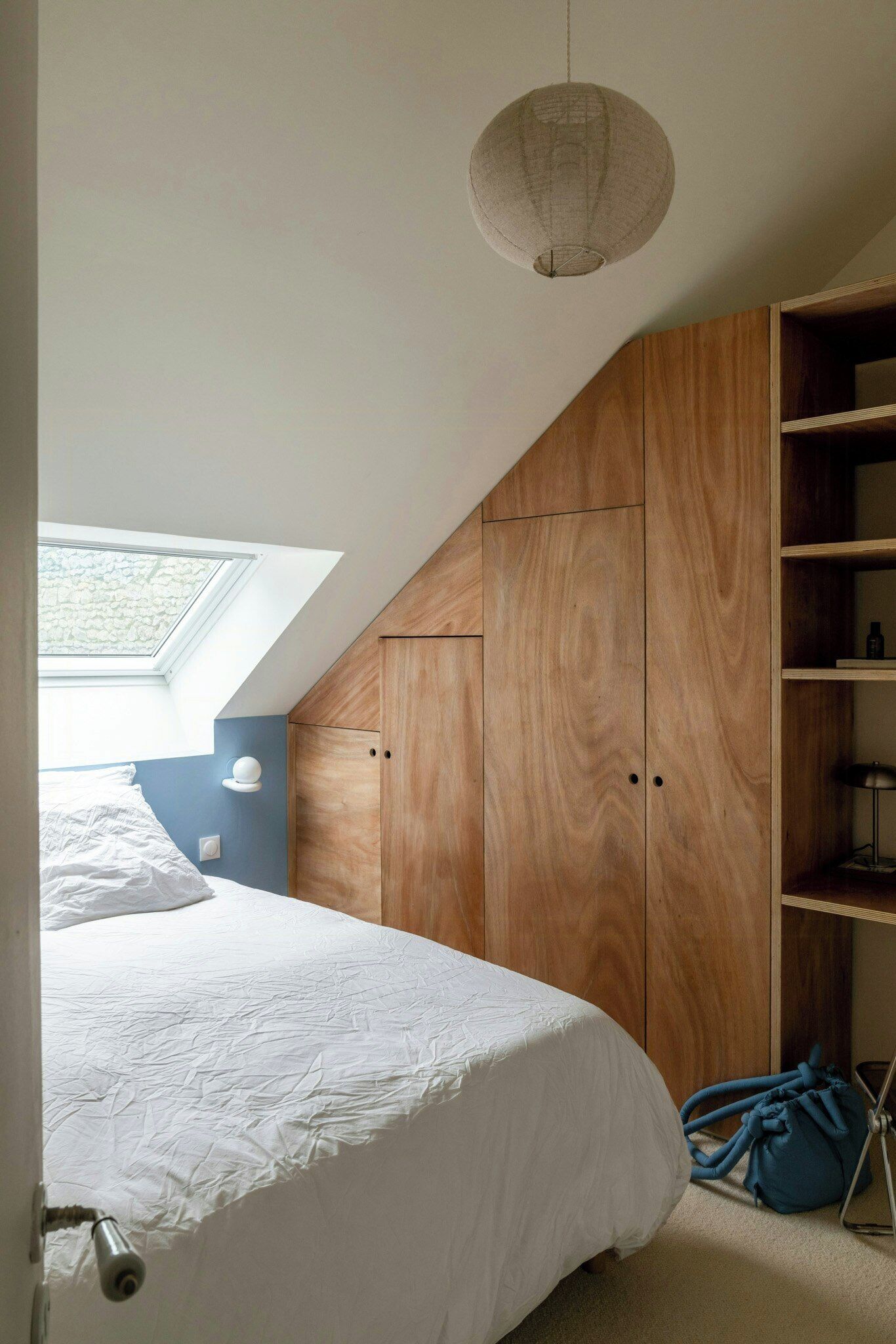 A bedroom with a skylight, a bed, wooden wardrobe, and a blue bag.