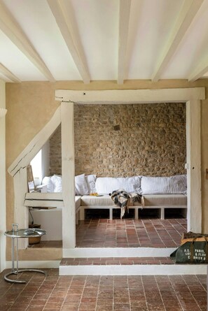 Ein rustikales Interieur mit einer Steinwand, einer weißen Bank mit Kissen, einem kleinen Tisch mit einem Korb und einer Tasche auf dem Boden.