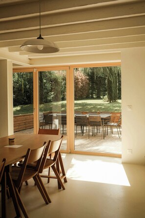Ein moderner Essbereich mit einem großen Tisch und Stühlen, einer Deckenleuchte und Blick auf einen Garten durch Glastüren.