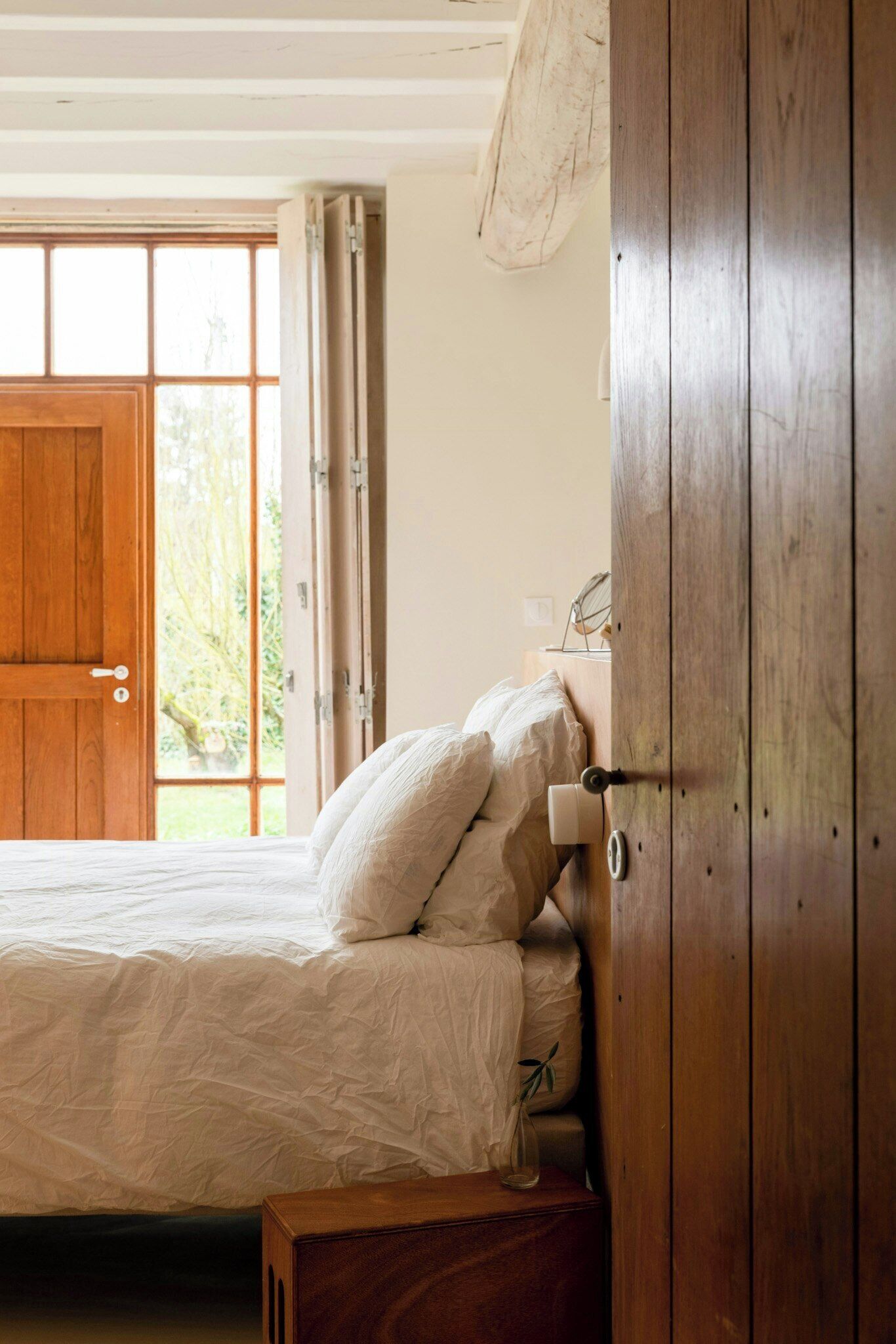 A bedroom with a wooden door, a bed with white bedding, and a window with a view of greenery.