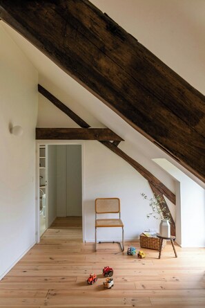 Ein Dachzimmer mit freiliegenden Holzbalken, ein Korbsessel, ein kleiner Tisch mit einer Vase und Spielzeug auf dem Boden.