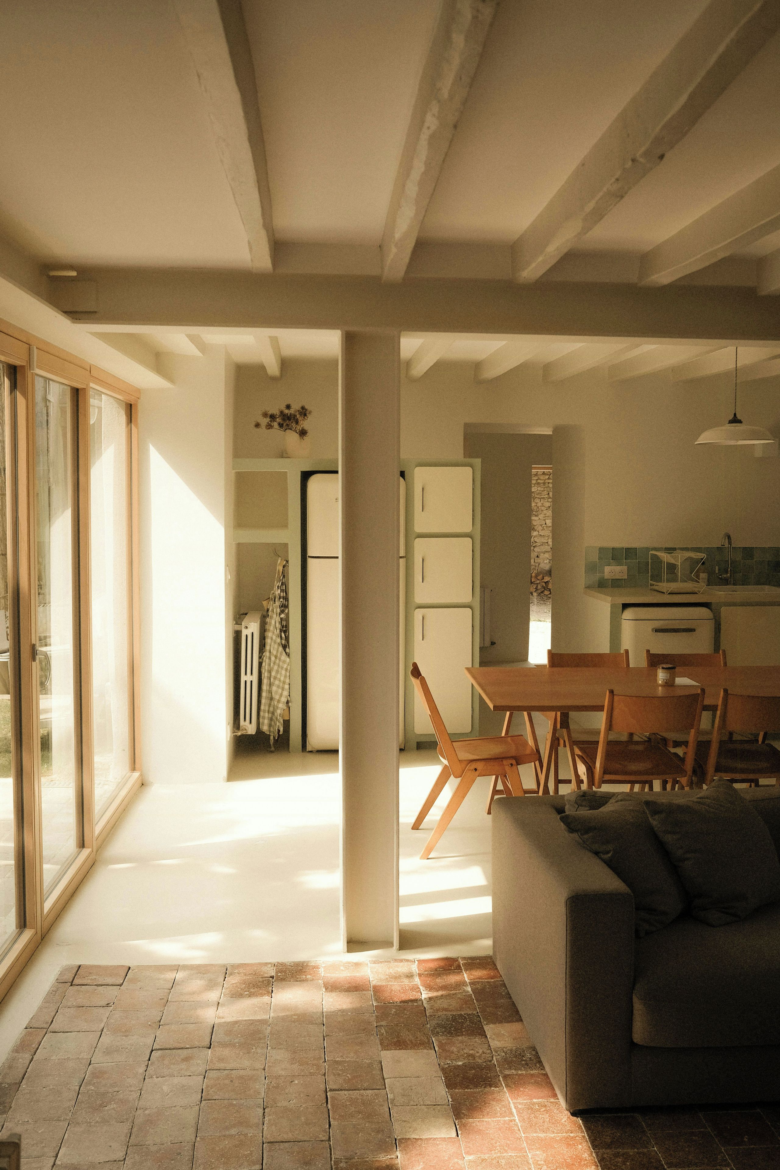 A spacious living area with a large window, a dining table with chairs, a sofa, and a kitchen in the background.
