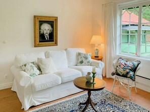 Ein weißes Sofa mit dekorativen Kissen, ein kleiner runder Tisch mit Kerze, ein Sessel mit Blumenkissen und ein gerahmtes Porträt an der Wand.