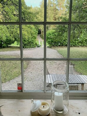 Ein Fenster mit Blick auf einen Kiesweg, der zu einer Holzterrasse führt, ein Glas mit einer Kerze und eine kleine weiße Kerze auf einer Holztischplatte.