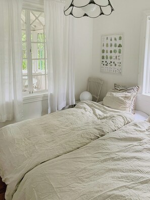 Ein Schlafzimmer mit einem großen Bett, einem Fenster mit Vorhängen, einer Wandleuchte und einem gerahmten Pflanzenbild.