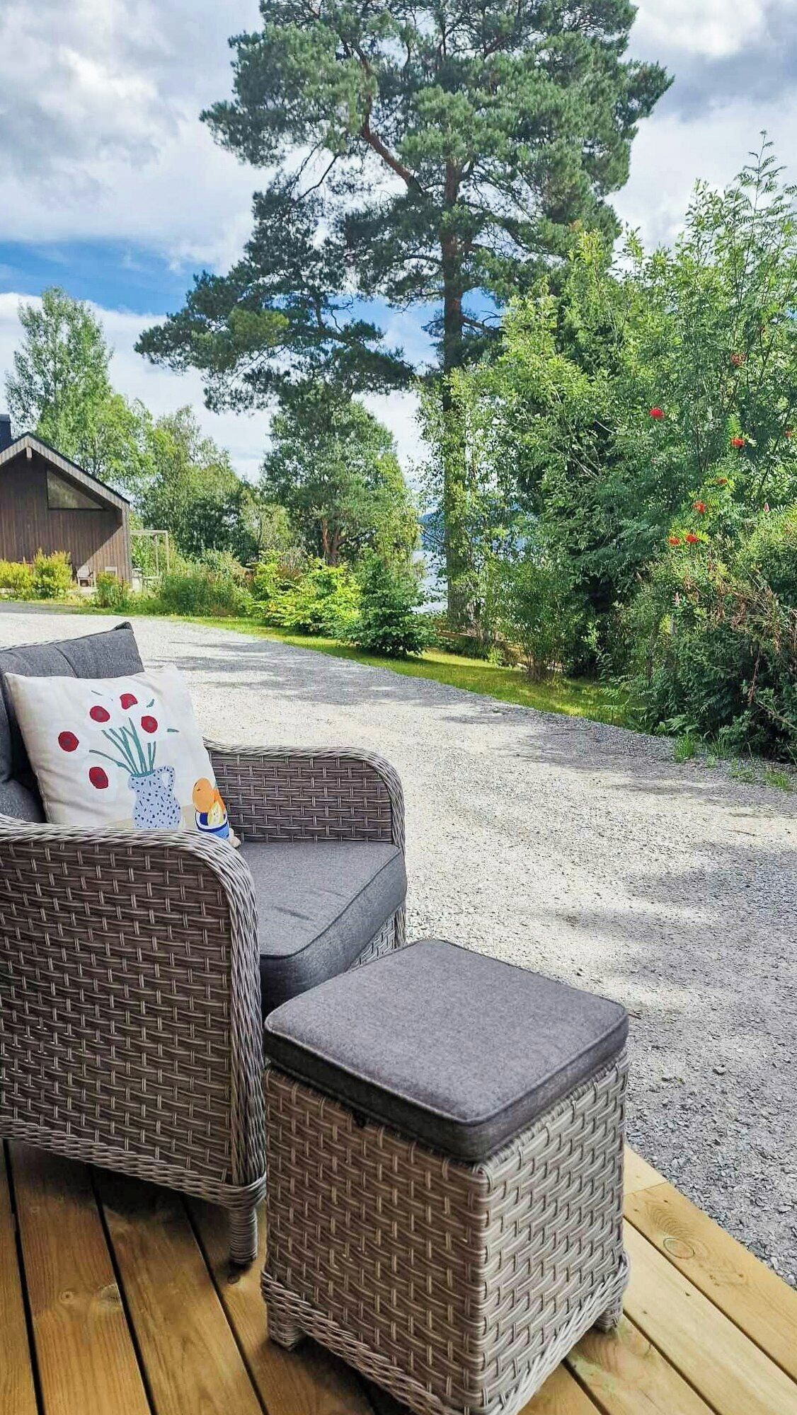 Un canapé et un pouf en osier, installés sur une terrasse en bois, avec un chemin de gravier menant à une maison.
