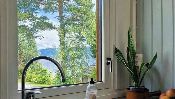 A kitchen with a double sink, a window with a view of trees and a body of water, a potted plant, and a bottle of hand soap. - Charming seaside cabin in Tingvoll (Tingvoll)