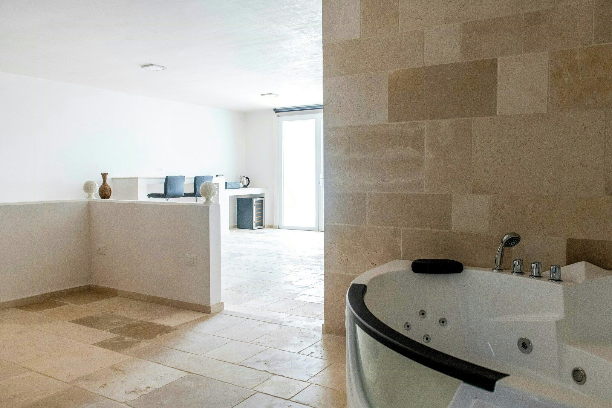 A modern bathroom with a large bathtub, a shower, and a toilet. There is a visible sink area with a mirror and a vase. The floor is tiled, and the walls are plain.