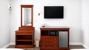 A hotel room with a wooden dresser, a microwave, a refrigerator, a TV, a mirror, and a trash bin.