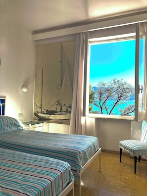 A room with a bed, a chair, a window with a view of the sea, and a sailboat painting on the wall.