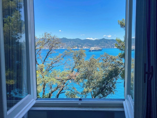 A view through a window of a coastal landscape with trees, a clear blue sea, and distant mountains.