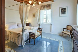 Ein Schlafzimmer mit einem Himmelbett, einer hölzernen Bank, einem Stuhl, einem Schminktisch und einem Fenster mit Vorhängen.