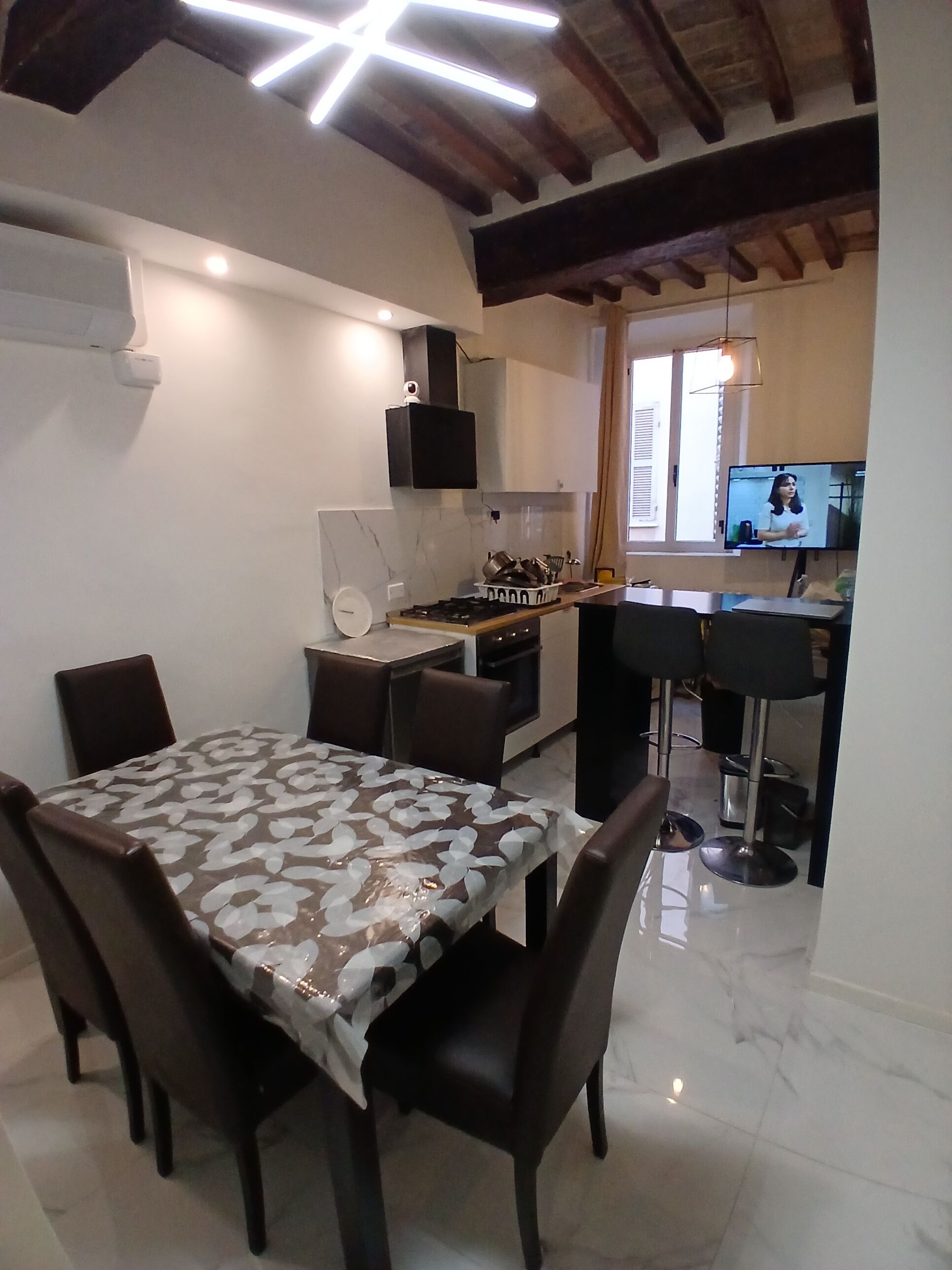 Un comedor con una mesa cubierta por un mantel estampado, rodeada de sillas. Se ve una cocina al fondo con electrodomésticos modernos y un televisor de pantalla plana sobre la encimera.