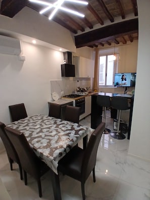 A dining area with a table covered in a patterned tablecloth, surrounded by chairs. A kitchen is visible in the background with modern appliances and a flat-screen TV mounted above the counter.