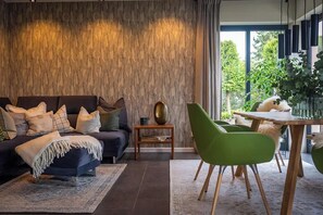 Ein modernes Wohnzimmer mit einem dunklen Sofa, grünen Sesseln, einem Holztisch und Blick ins Grüne.