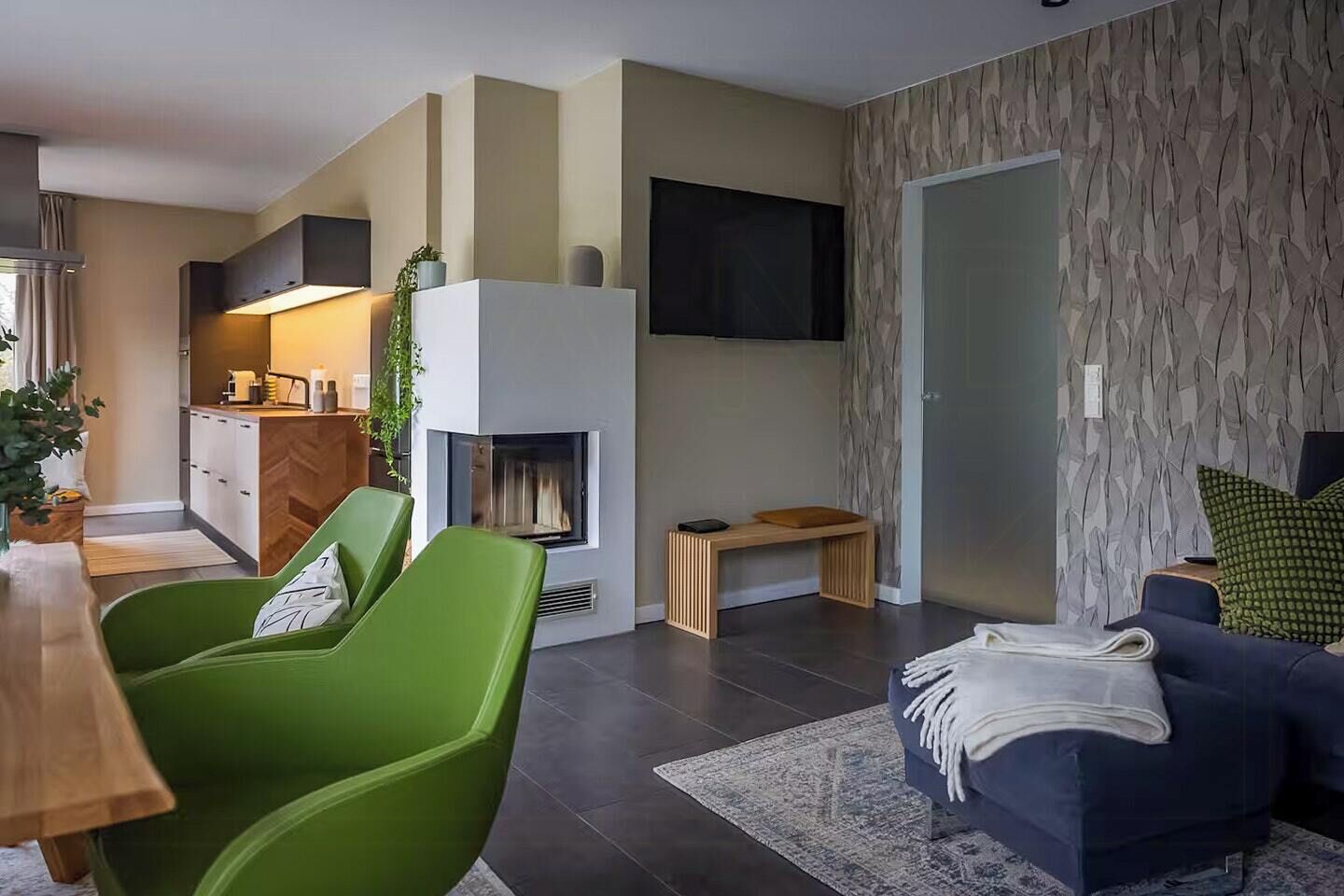 A modern living room with a fireplace, green chairs, a dark sofa, and a kitchen area.
