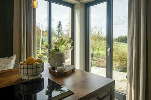 Ein Zimmer mit großem Fenster, einem Holztisch, einer Obstschale, einer Blumenvase und einer Kerze.