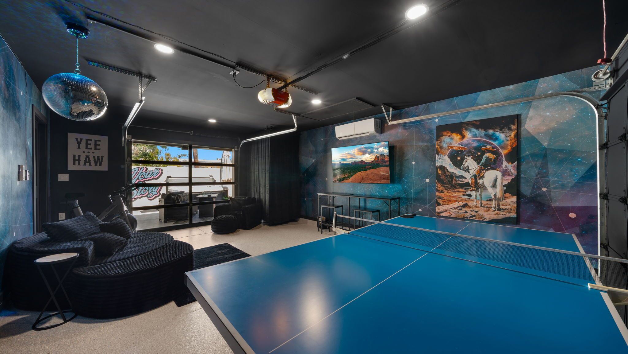 A room with a ping pong table, black furniture, a disco ball, and a mural with a space theme.