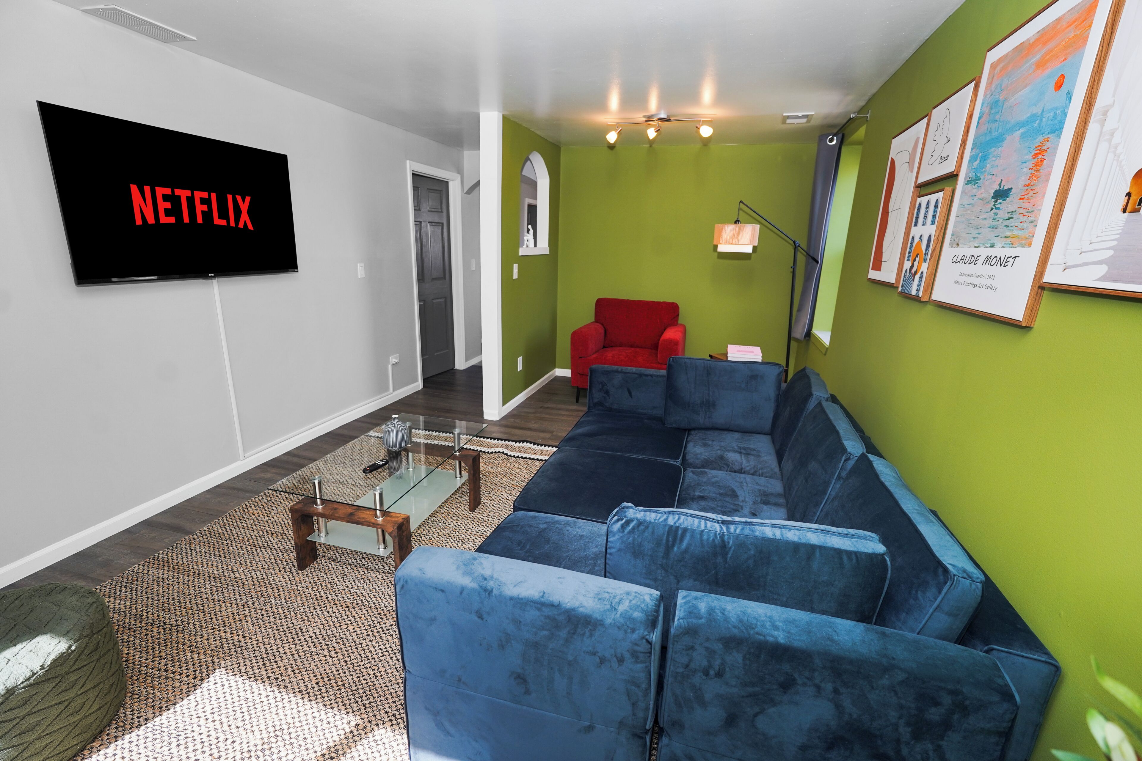 Ein Wohnzimmer mit einem großen Flachbildfernseher, der 'NETFLIX' anzeigt, einem blauen Ecksofa, einem roten Sessel, einem Glastisch und gerahmten Kunstwerken an den Wänden.