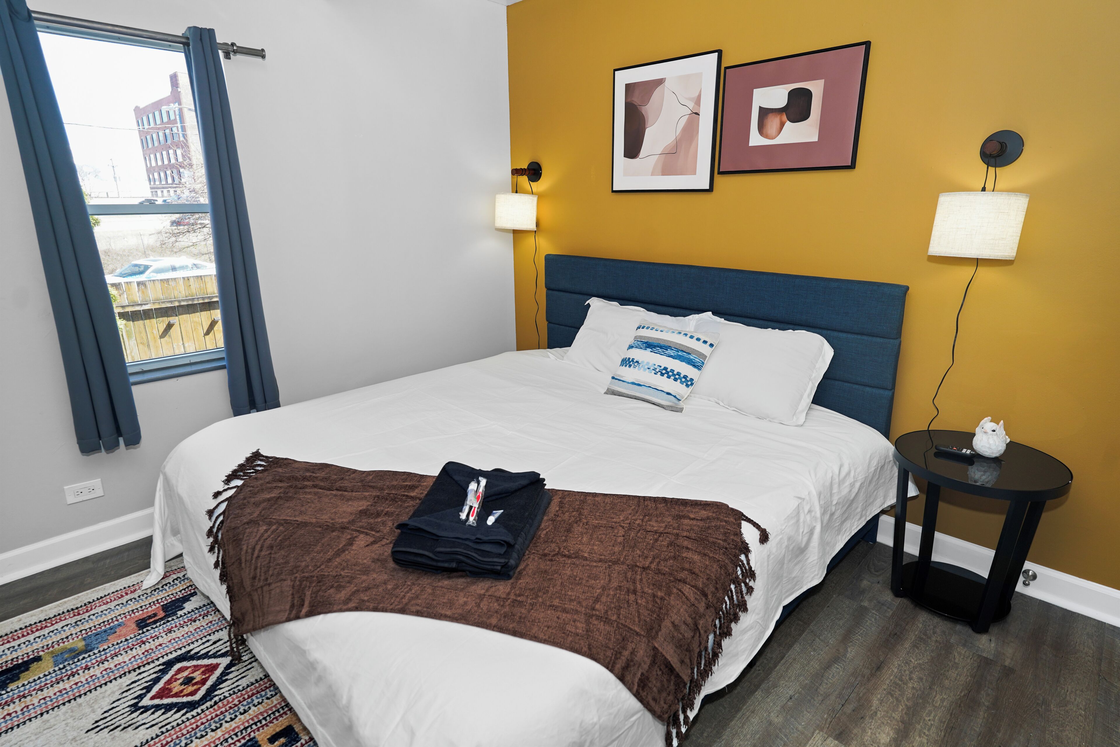 A hotel room with a bed, a small side table, a wall-mounted lamp, and two framed pictures.