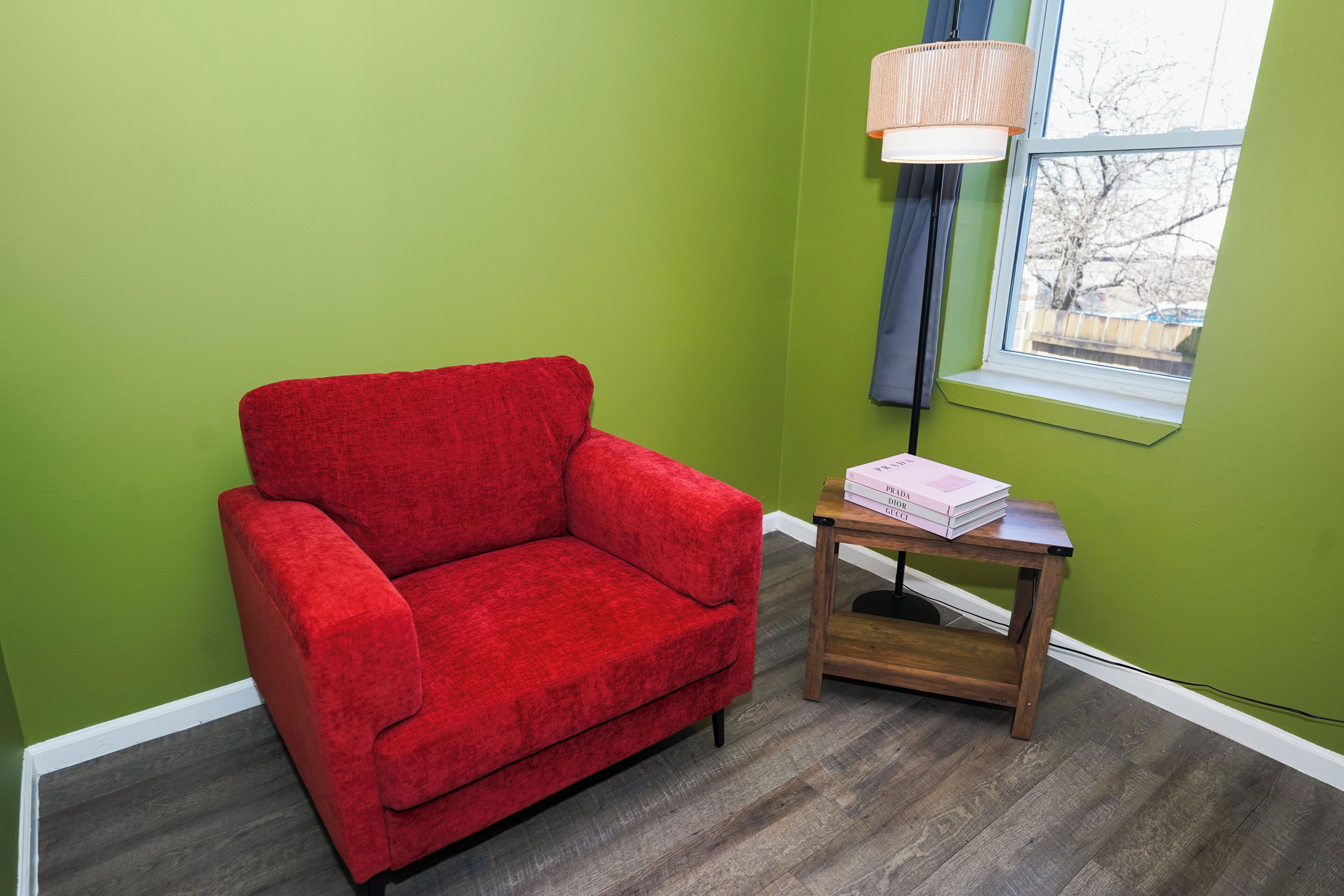 Un fauteuil rouge, une petite table en bois avec des livres, une lampe et une fenêtre donnant sur un beau paysage.