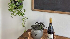 Uma mesa de madeira com uma garrafa de vinho, uma planta em vaso e uma lousa numa parede branca.