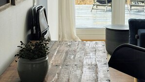 Uma mesa de madeira com um vaso de planta, uma cadeira preta e vista para a praia através de um vão de vidro.