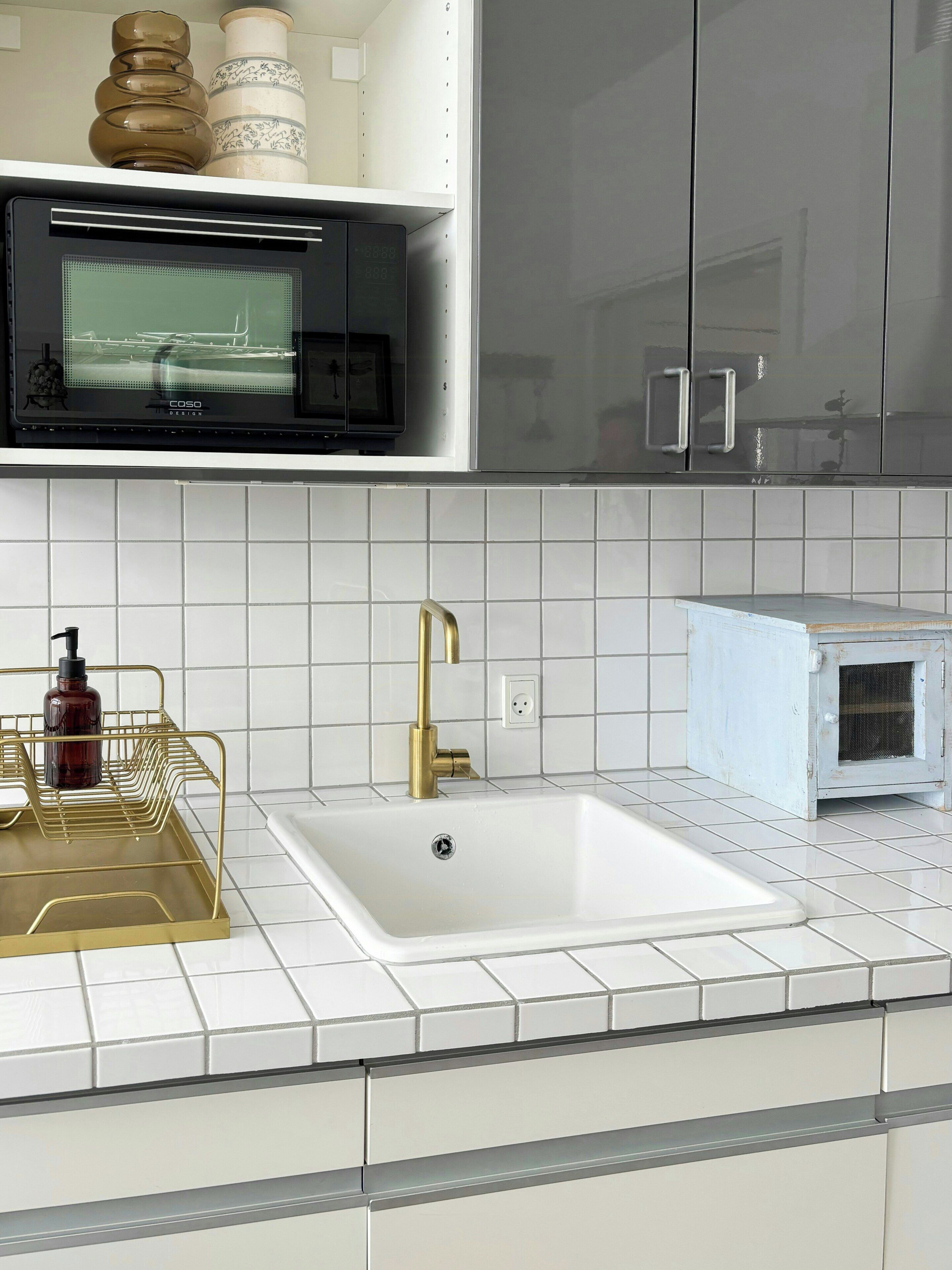 A kitchen with white tiled backsplash, a white sink, a gold faucet, and a small white appliance on the countertop.