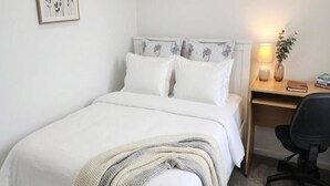 A neatly made bed with white bedding and a beige throw blanket, a wooden desk with a lamp and a book, and a black office chair. - Stylish Wesley Retreat (Auckland)