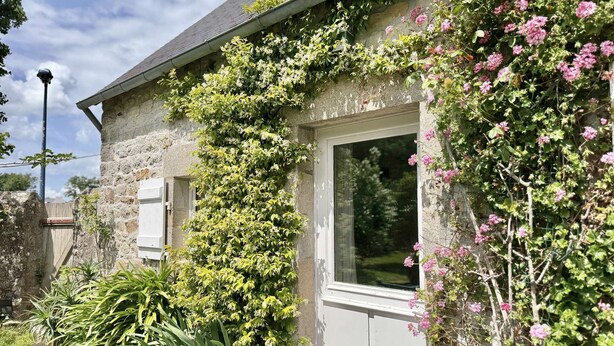A stone building with a white door and window, surrounded by lush greenery and flowering plants. - Bénodet, charming stone house with beautiful garden (BENODET)