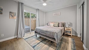 A bedroom with a bed, bedside tables, a nightstand, a ceiling fan, and a window with curtains. - Checkmate Cottage - Fire Pit + Putting Green (Palm Harbor)