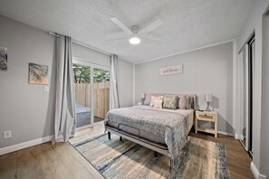A bedroom with a bed, bedside tables, a nightstand, a ceiling fan, and a window with curtains. - Checkmate Cottage - Fire Pit + Putting Green (Palm Harbor)