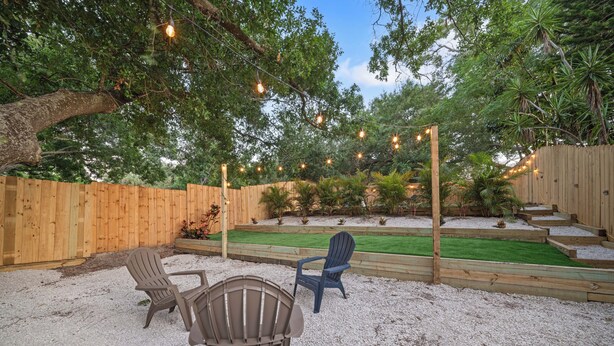 A backyard with a wooden fence, two Adirondack chairs, a grassy area, and string lights. - Checkmate Cottage - Fire Pit + Putting Green (Palm Harbor)