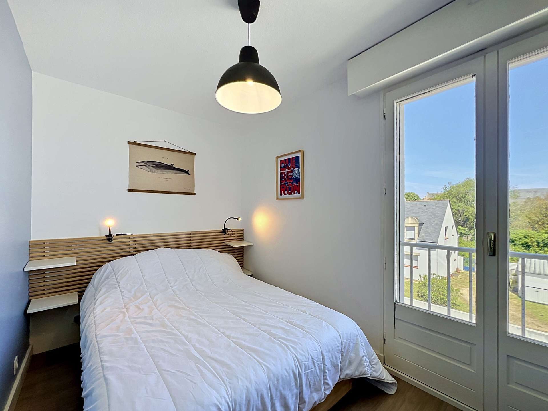 A bedroom with a bed, a headboard, a wall-mounted lamp, a framed picture, and a window with a view.