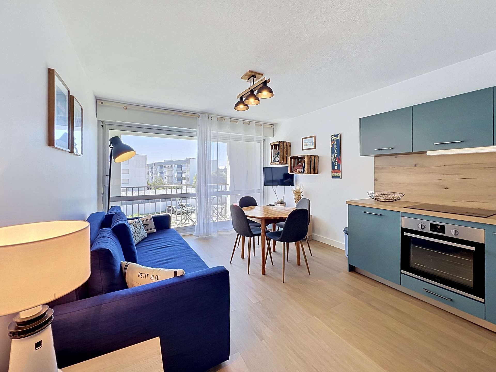 A modern apartment with a blue sofa, dining table, chairs, a kitchen with teal cabinets, and a large window.