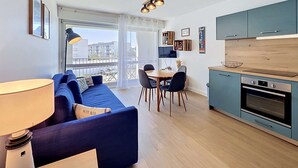 A modern apartment with a blue sofa, dining table, chairs, a kitchen with teal cabinets, and a large window. - QUIBERON - 2 rooms - 33m2 - City center (QUIBERON)