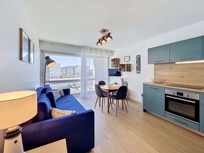 A modern apartment with a blue sofa, dining table, chairs, a kitchen with teal cabinets, and a large window. - QUIBERON - 2 rooms - 33m2 - City center (QUIBERON)