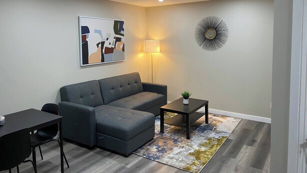 A modern living room with a grey sectional sofa, a small black table with a plant, a floor lamp, and a wall art piece. - Serene and Stylish House Rental in Brighton, Saskatoon (Saskatoon)