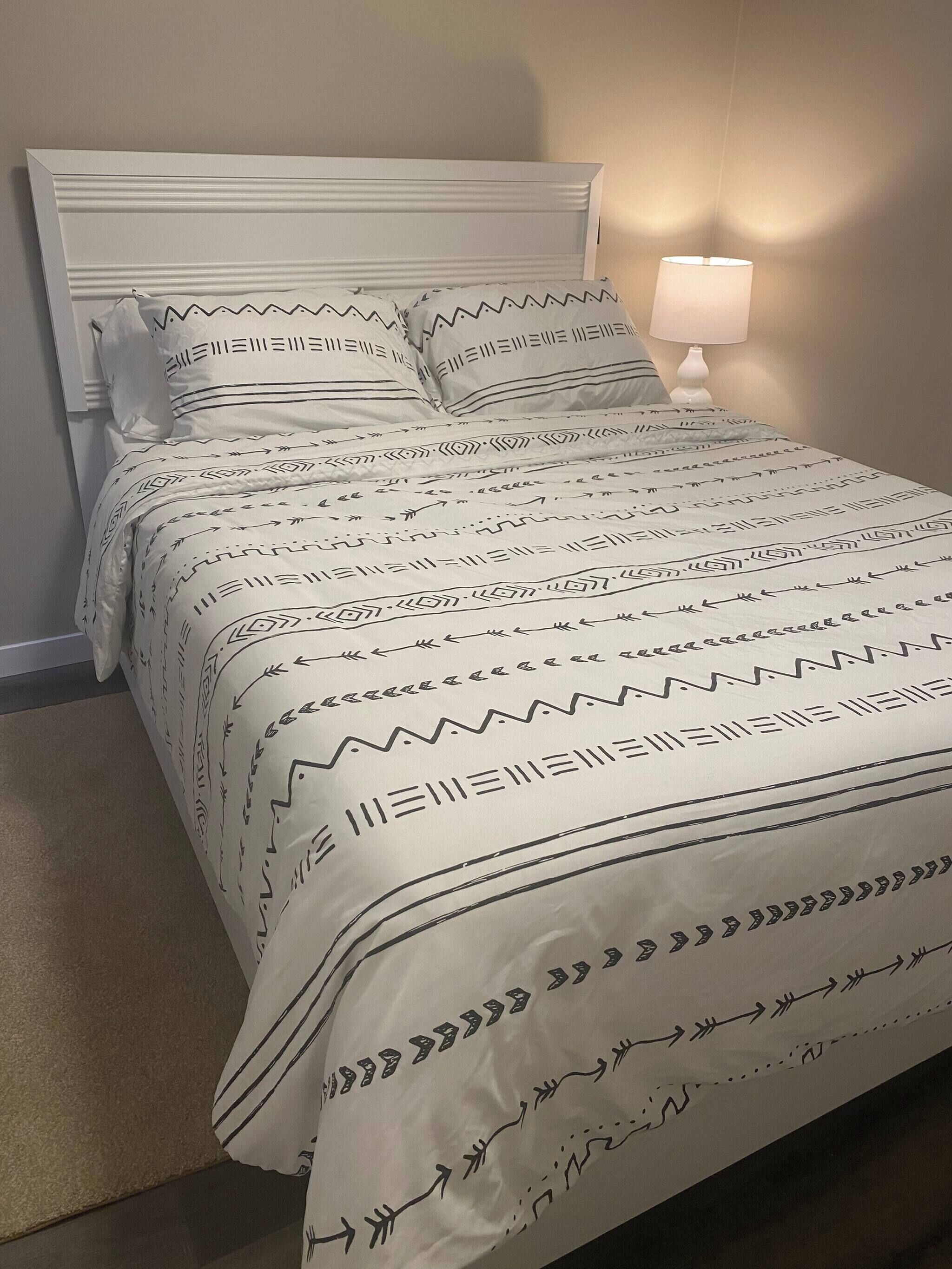 A bed with a patterned bedspread and pillows, a bedside lamp, and a plain wall.