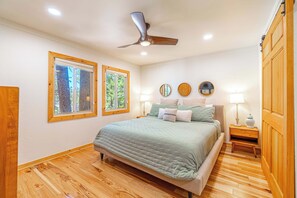 Ein Schlafzimmer mit einem Bett, Holzboden, Deckenventilator, zwei Fenstern und einem Nachttisch mit Lampe.