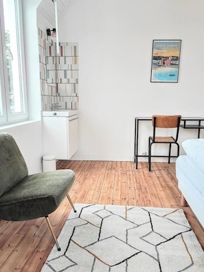 Een moderne kamer met een houten vloer, een groene stoel, een wit bed, een klein tafeltje met een stoel, een witte kast en een lijst met een afbeelding aan de muur.