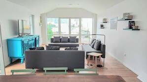 A modern living room with a sofa, a coffee table, and a TV. There are bookshelves and a window with a view.