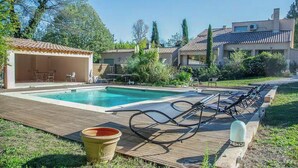 Une piscine avec des chaises longues, un patio couvert et un jardin agrémenté d’arbres et d’arbustes.