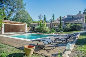 Ein Schwimmbad mit Liegestühlen, eine überdachte Terrasse und ein Garten mit Bäumen und Sträuchern.