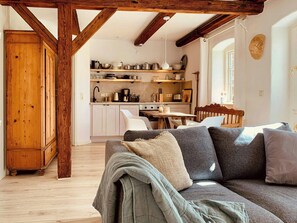 Ein gemütliches Wohnzimmer mit Holzbalkendecke, einer Couch mit Kissen, einem Esstisch und einer Küchenzeile mit Schränken und Geräten.