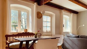 Een gezellige kamer met een houten eettafel, witte stoelen, een grijze bank en grote ramen met gordijnen.