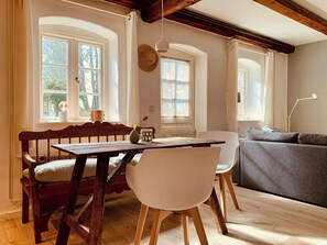 Ein gemütliches Zimmer mit Holztisch, weißen Stühlen, einem grauen Sofa und großen Fenstern mit Vorhängen.