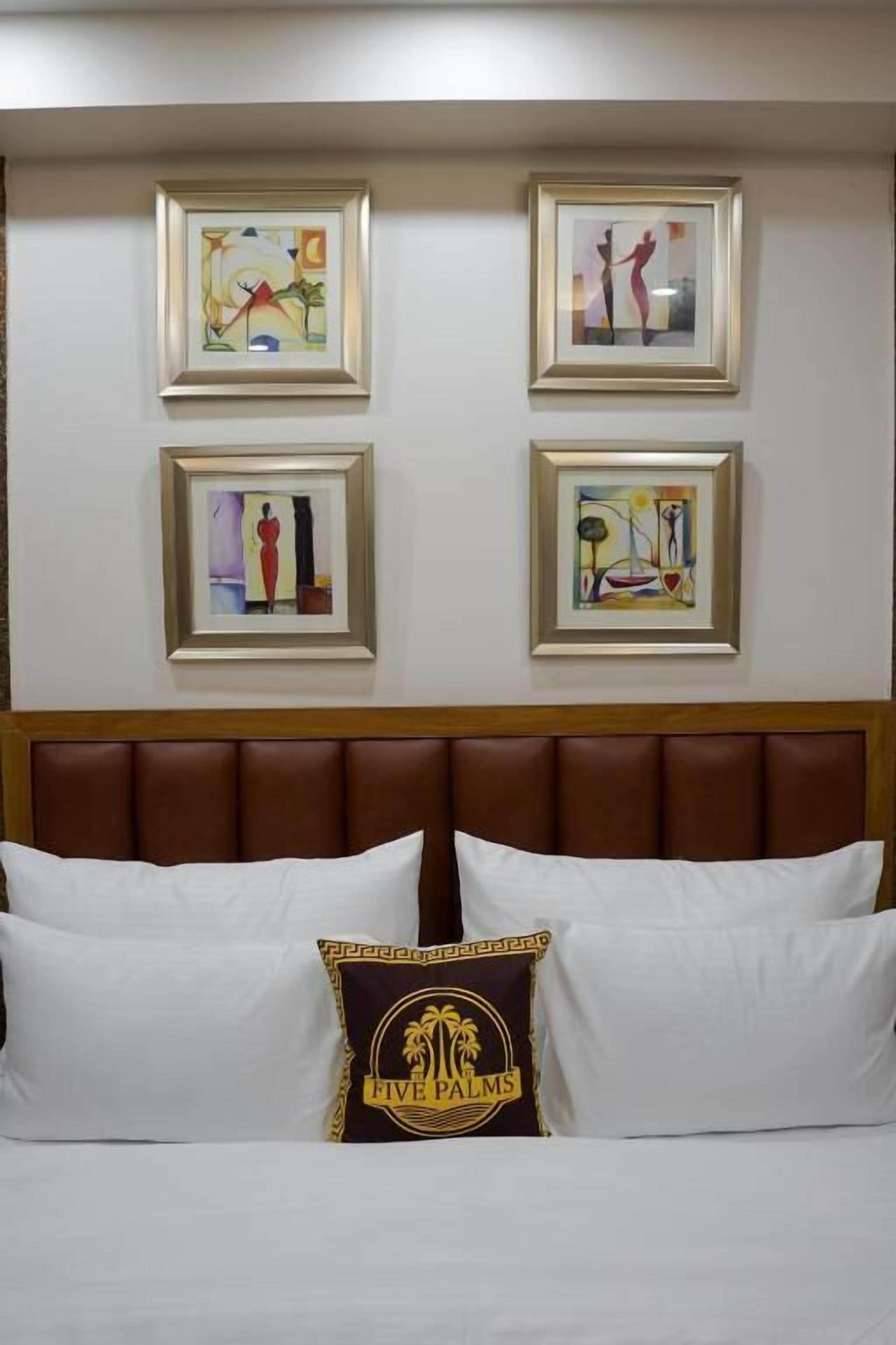 A hotel room with a bed, white pillows, and a headboard. There are four framed artworks on the wall.