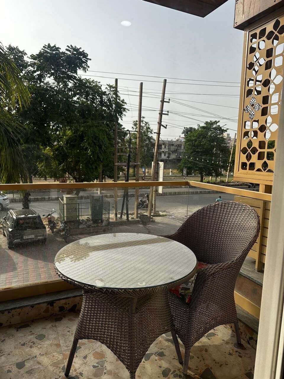 A balcony with a wicker table and chairs, overlooking a street with vehicles and trees.